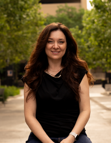 A woman with long brown hair wearing a black top, smiling outdoors with trees and buildings in the background.