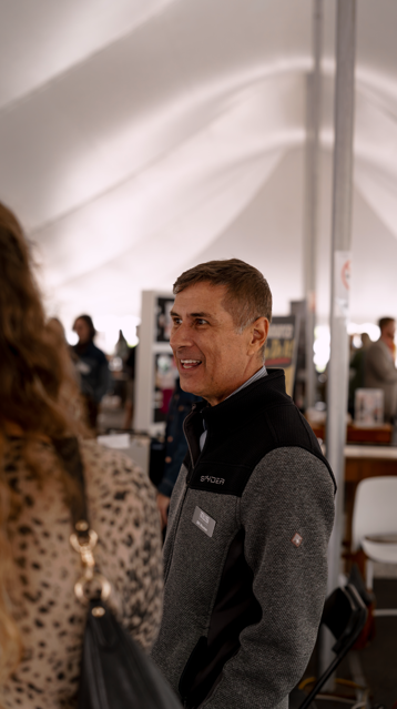 A man smiling and talking to a woman in an indoor event or expo with booths and other people in the background.