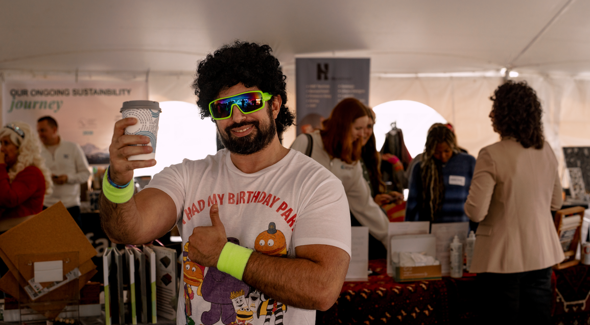A man with curly hair and a beard wearing colorful sunglasses, a white T-shirt with a birthday-themed design, and neon wristbands, smiling and giving a thumbs-up while holding a disposable coffee cup at an indoor event with multiple people in the bac