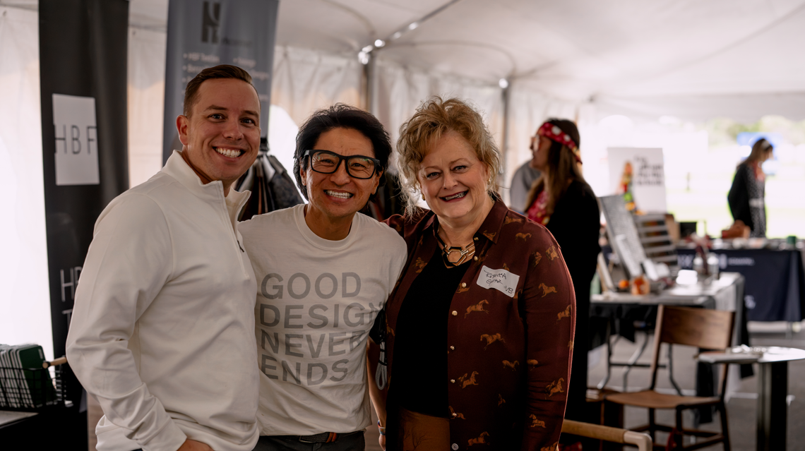 Three smiling people standing inside a tent, with a woman wearing a patterned blouse and others in the background, at an event or market.