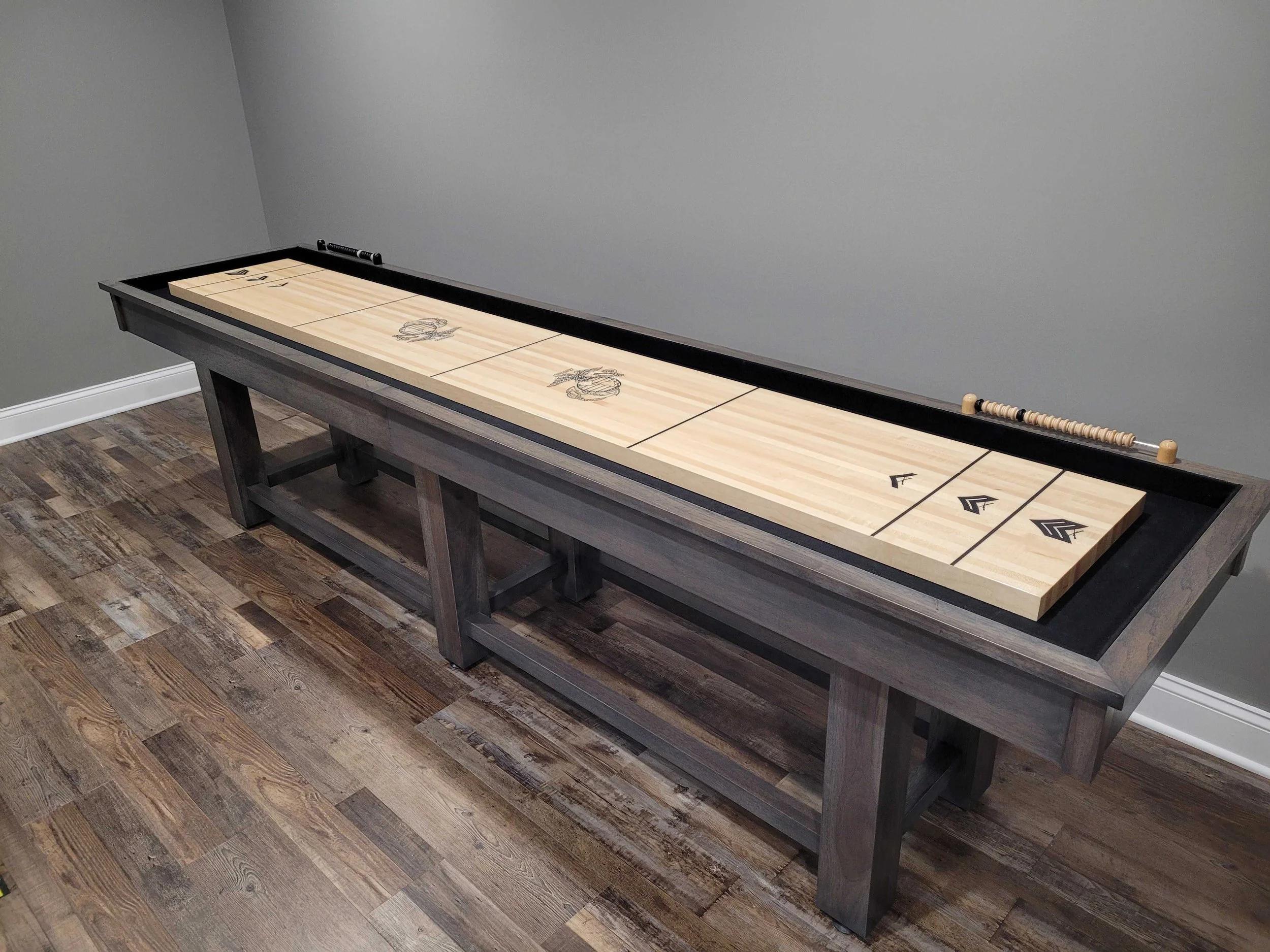 A wooden shuffleboard table with black edges, featuring a detailed eagle engravings and black arrow markings at one end, situated against a plain gray wall on a wood-patterned floor.