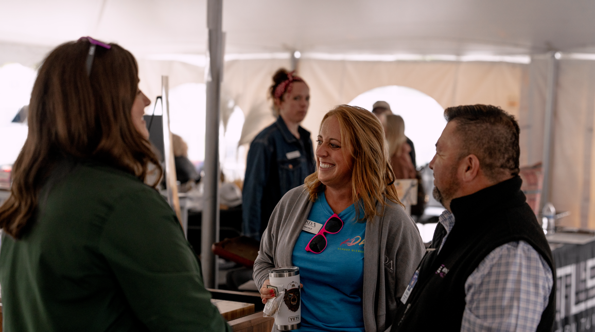 Three people smiling and talking inside a tent, with a woman in a blue shirt holding a YETI tumbler.