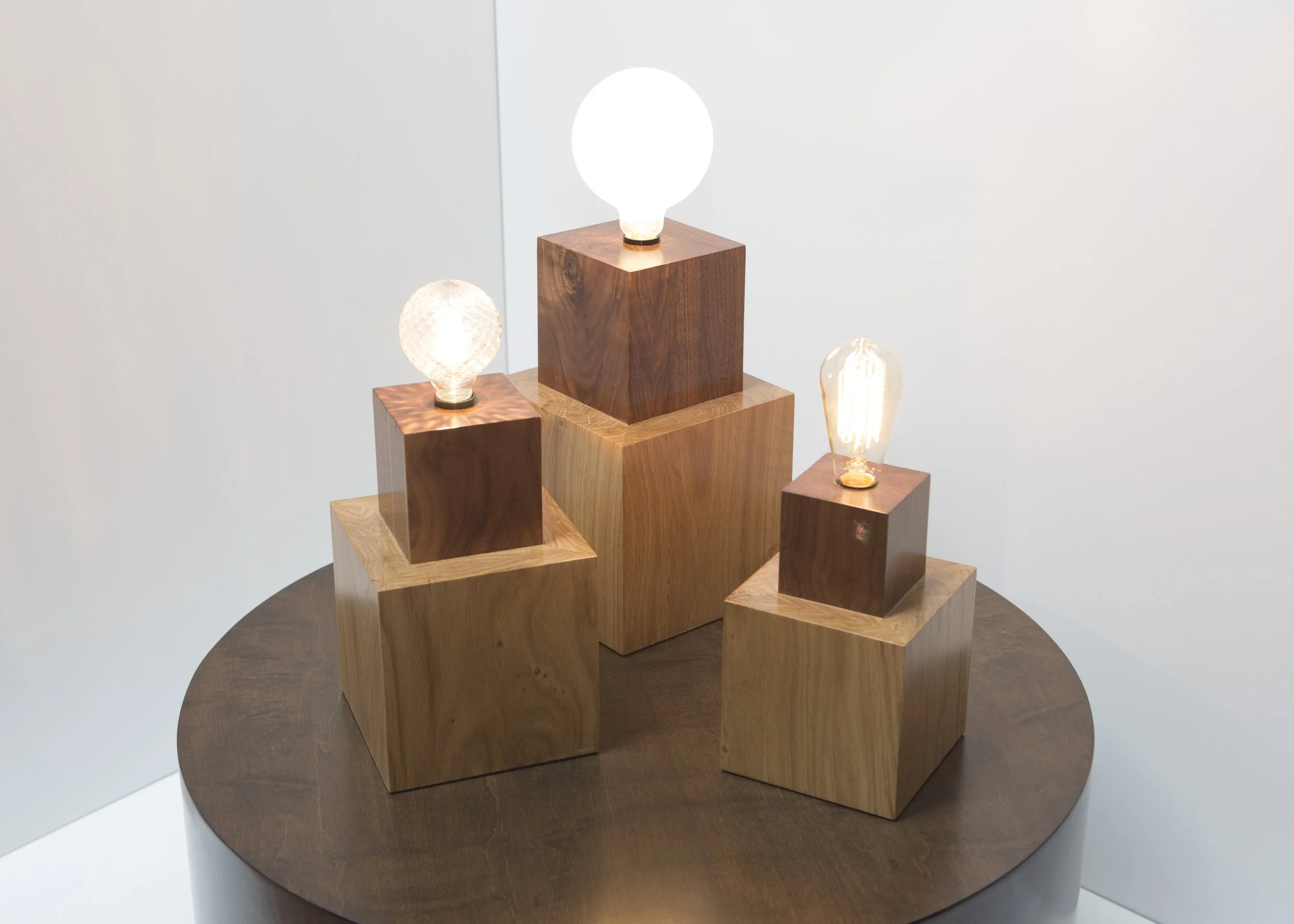 Display of four modern table lamps with different shaped bulbs on wooden pedestals, arranged on a round wooden table.