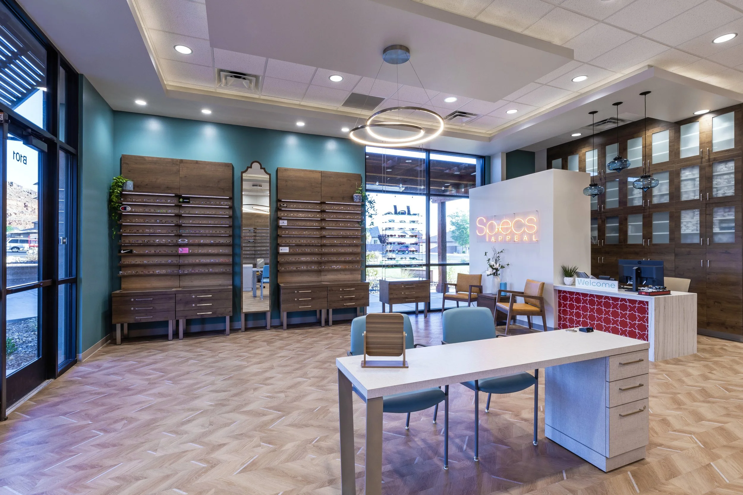 Interior of a modern eyeglasses store with display walls, a reception desk, and seating area, illuminated by ceiling lights and a circular hanging light fixture.