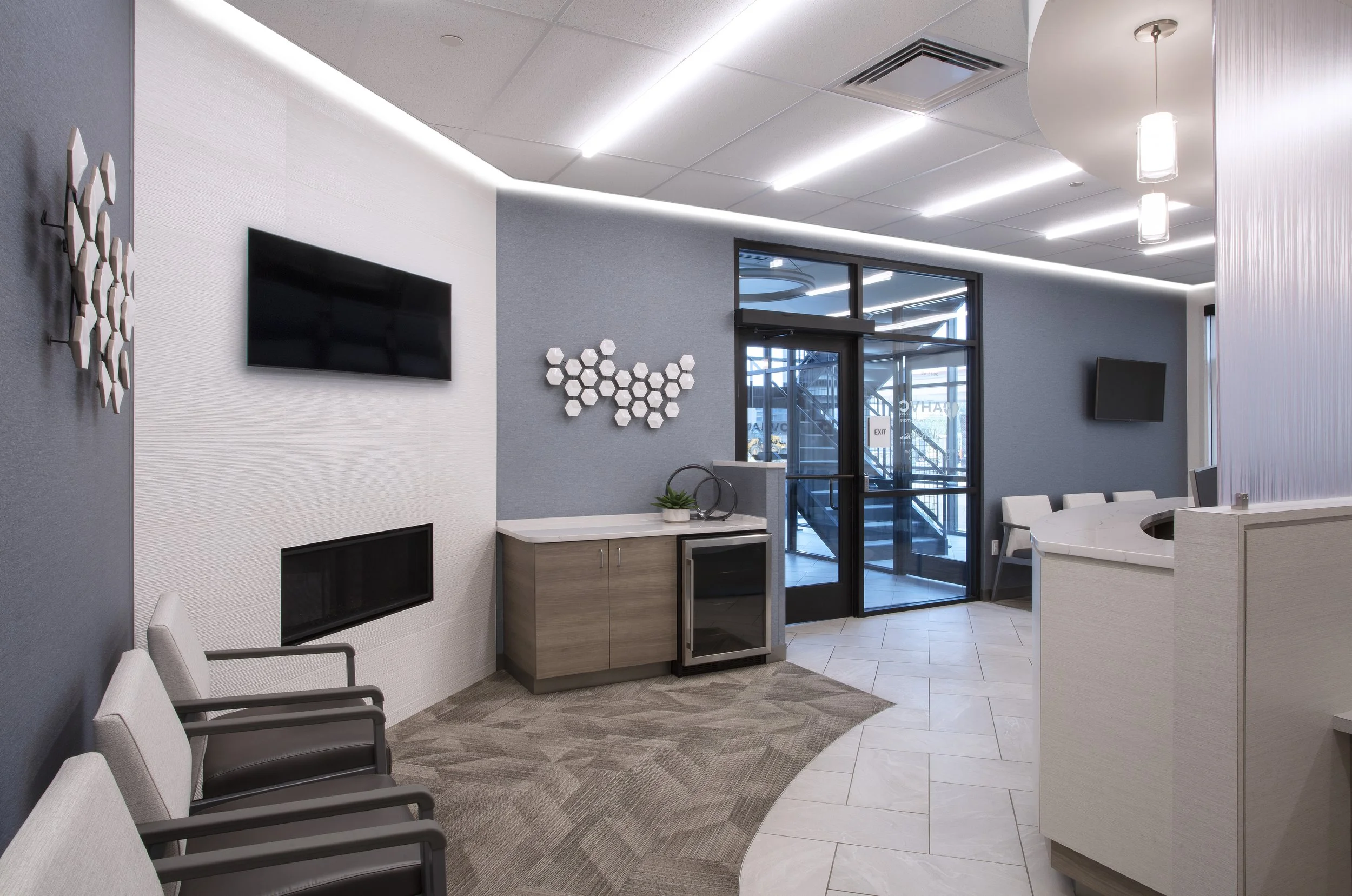 Modern lobby area with gray and white walls, wall-mounted flat-screen TVs, decorative white hexagonal wall art, a glass door entrance, and seating with white chairs along a curved counter.