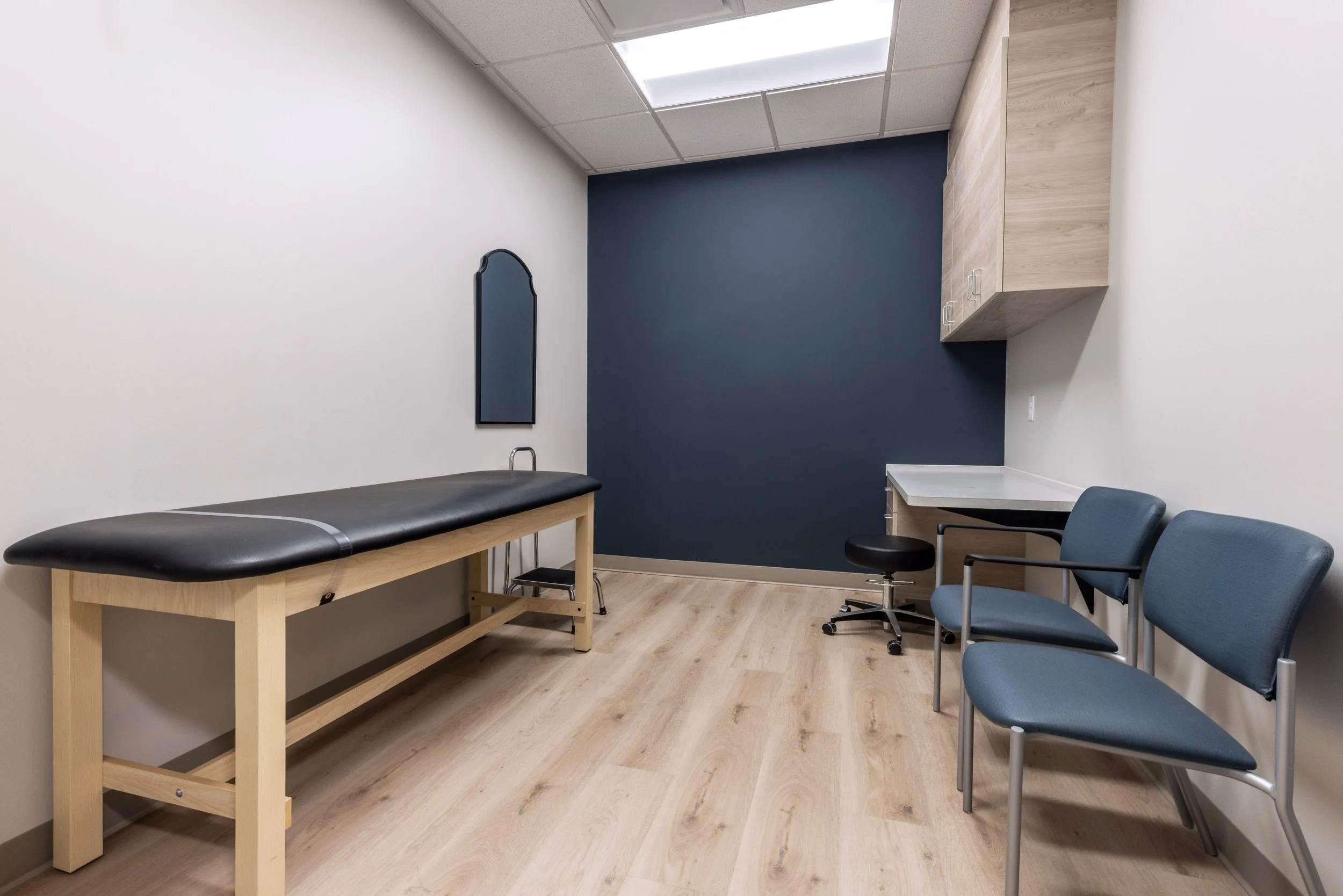 Medical examination room with a black examination table, blue chairs, a wooden cabinet, and a blue accent wall.