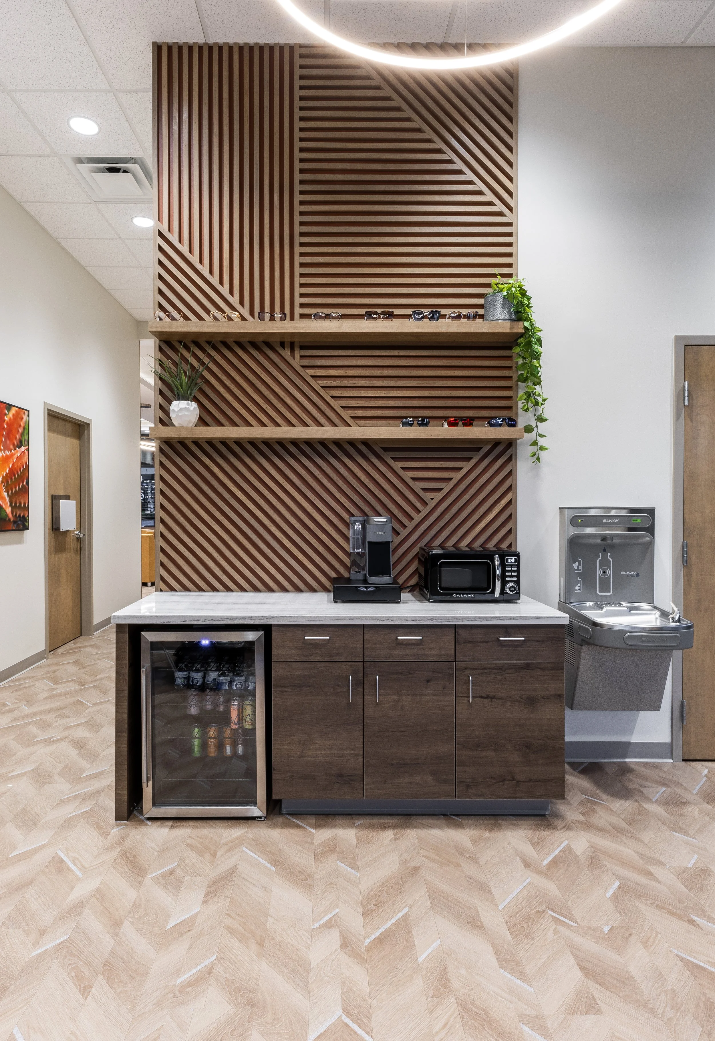 Coffee station with a white countertop, a coffee maker, microwave, small refrigerator, water dispenser, and decorative plants on wooden shelves with geometric slat design in the background.