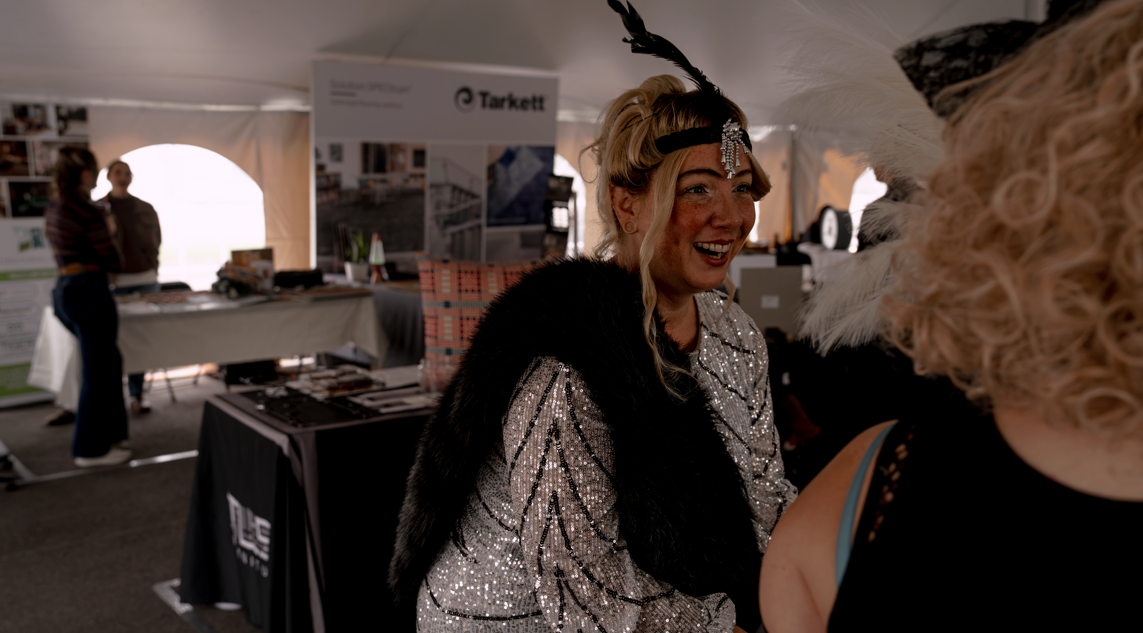 A woman wearing a shiny, patterned dress, a black fur stole, and a headpiece with feathers and jewels, smiling and talking to another person in a festival or event tent.
