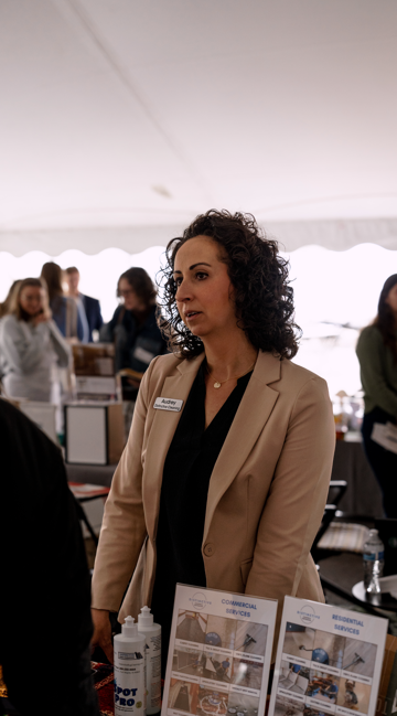 Woman with curly hair wearing a beige blazer at a booth with informational flyers, in a busy event setting.