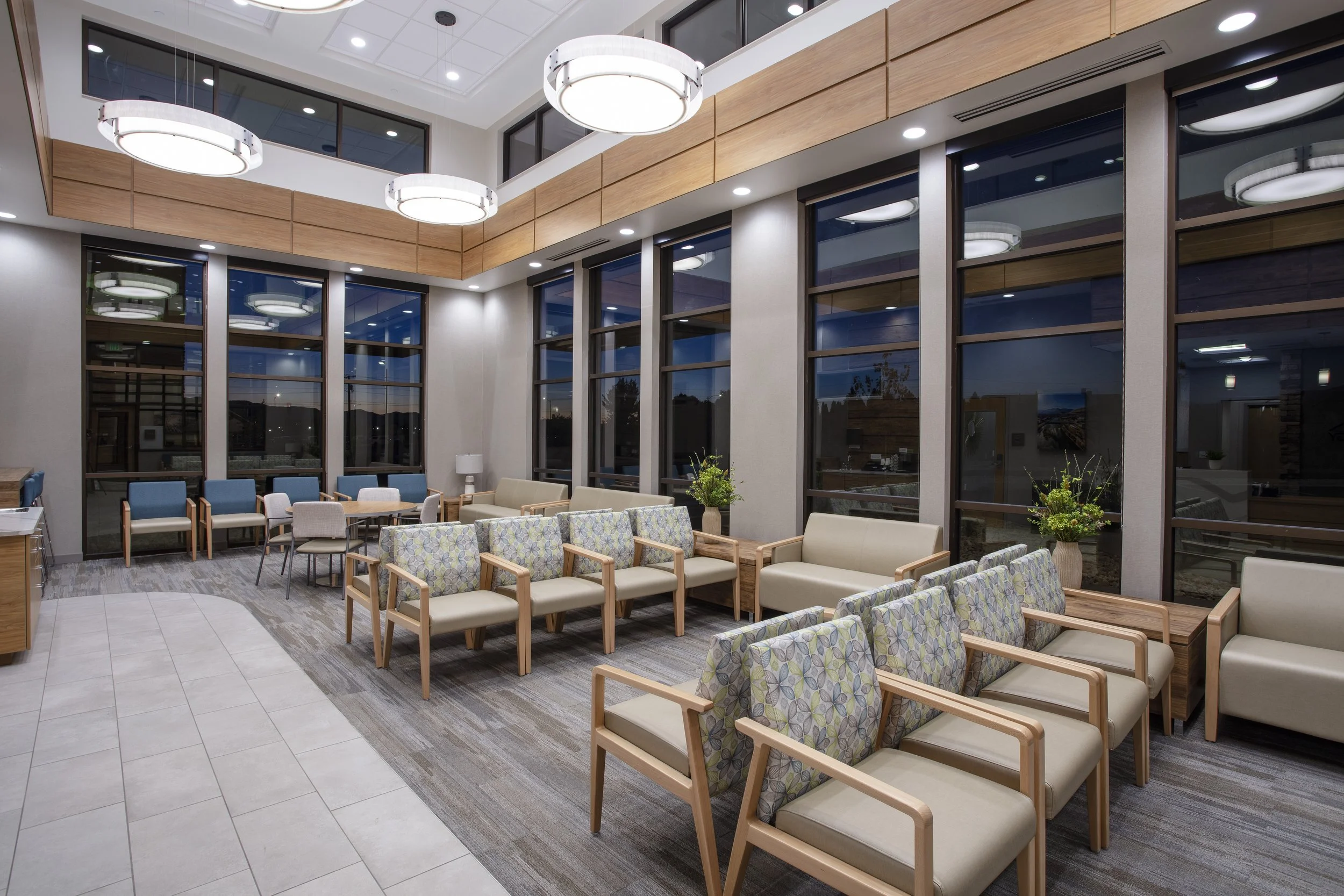 Indoor lounge area with multiple rows of beige and patterned chairs, large floor-to-ceiling windows, wood paneling, ceiling lights, and modern chandeliers.
