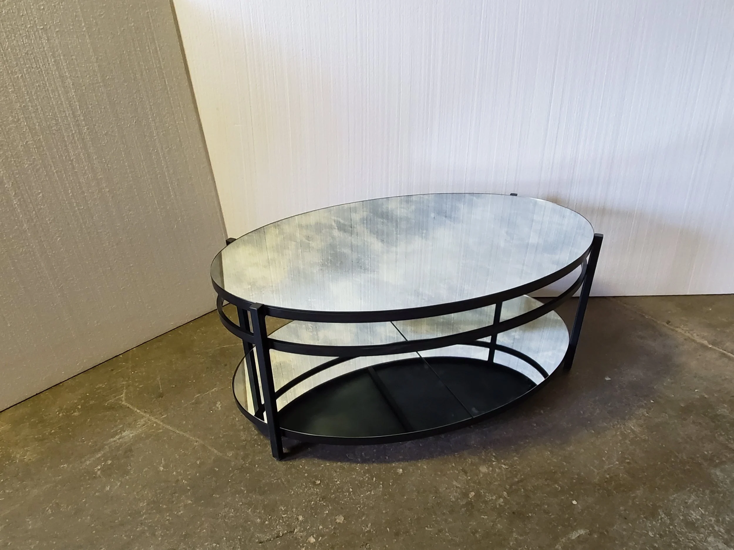 A round glass table with a black metal frame is placed indoors against beige and white textured walls. The floor appears to be made of a dark stone or tile.