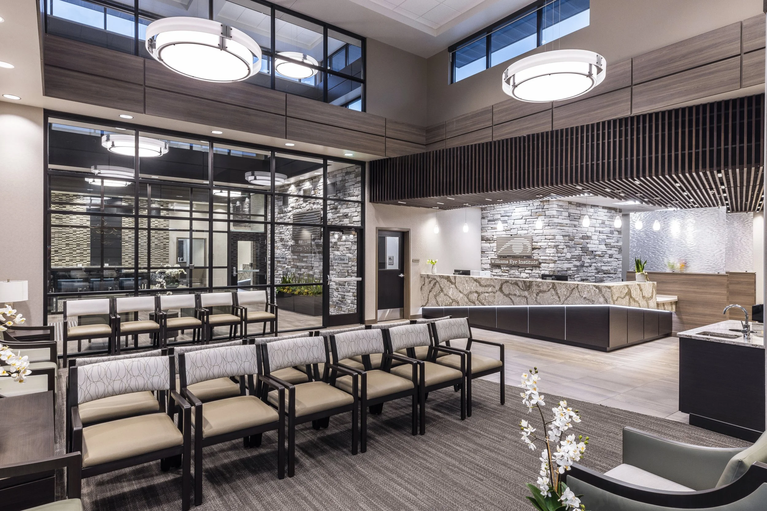 Interior view of a modern clinic waiting area with rows of beige chairs, a reception desk made of stone, and contemporary lighting fixtures, including large circular ceiling lights.