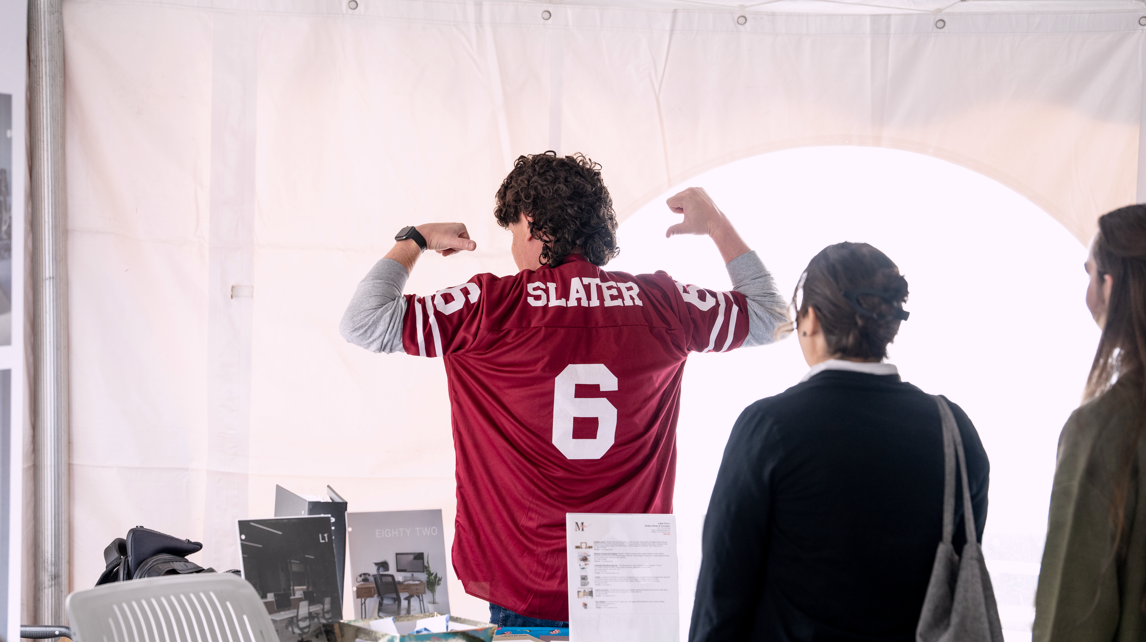 Person wearing a red sports jersey with the number 6 and the name Slater on the back, flexing muscles, with a group of three people watching, indoors near a table and window.