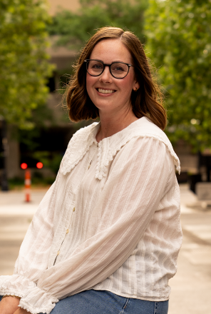 A woman with shoulder-length brown hair, glasses, and a white long-sleeve blouse smiling outdoors with trees and a city street in the background.