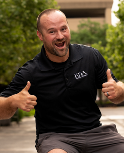 A smiling man with a shaved head and beard, giving two thumbs up while sitting outdoors, wearing a black polo shirt.