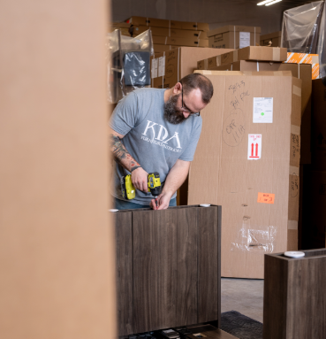 A man with tattoos and glasses assembling furniture in a warehouse surrounded by large cardboard boxes.