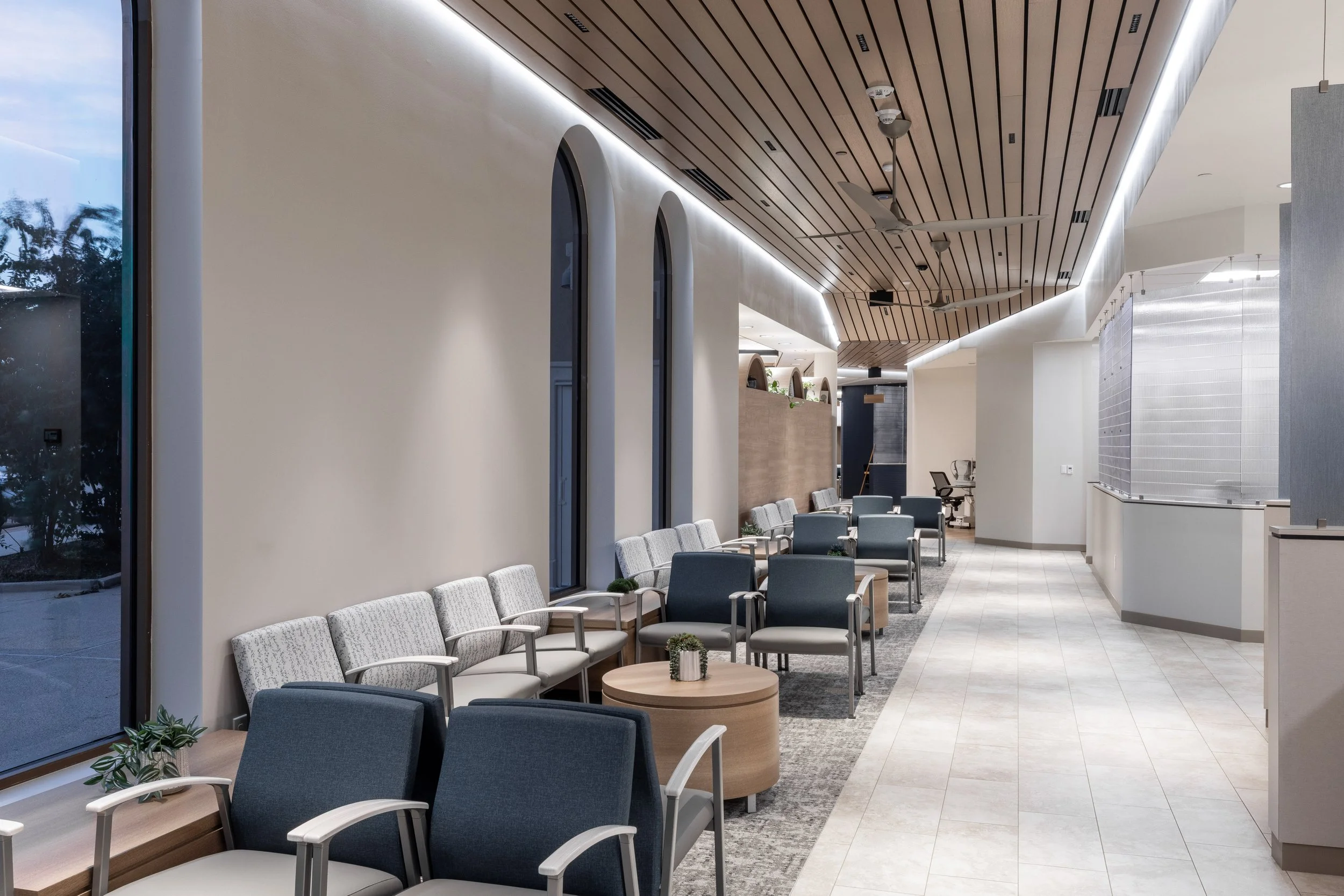 Modern interior of a waiting area or lobby with rows of chairs, large windows, and a wooden ceiling with recessed lighting and ceiling fans.