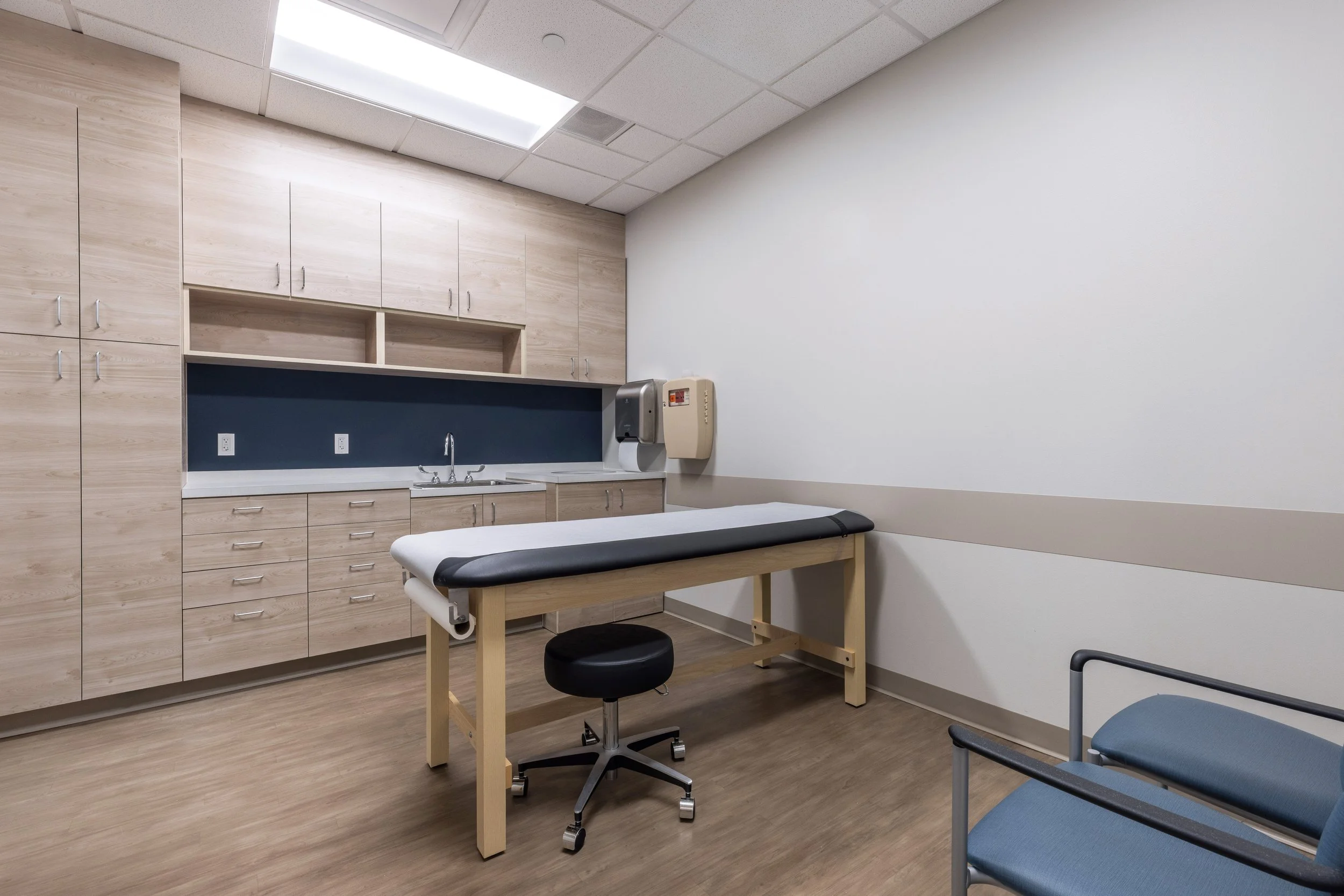 A medical examination room with wooden cabinets, a sink, a stool, and a patient chair. The room has a ceiling with fluorescent lights and light-colored walls.
