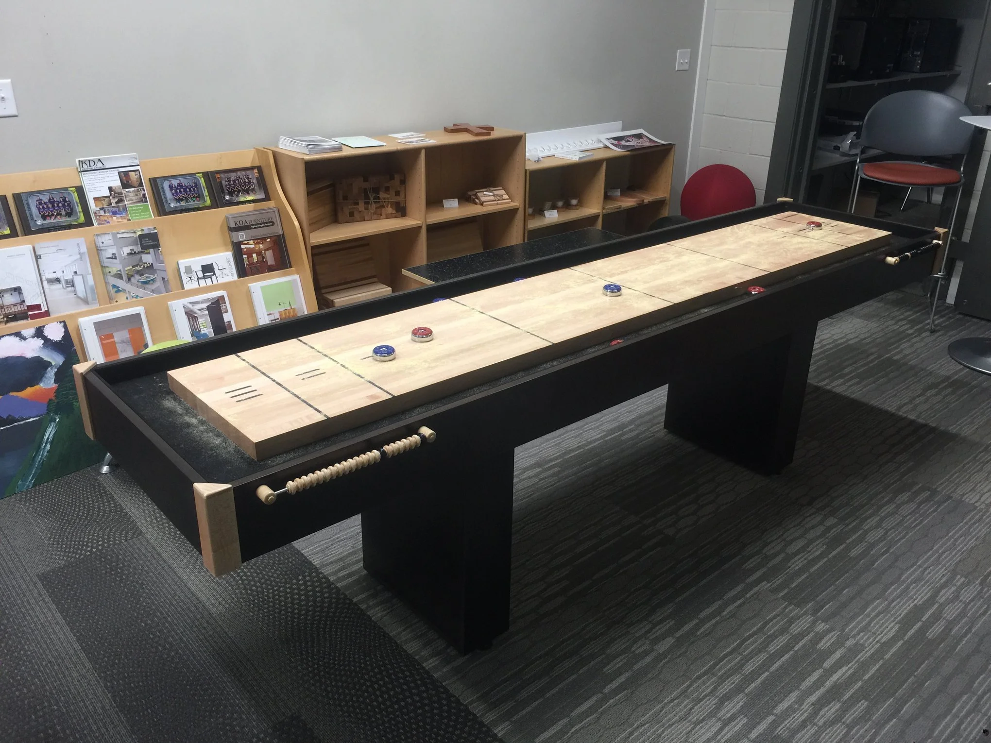 A shuffleboard game with pucks on a black and wood surface in an office or recreational room, with shelves of magazines and a red chair in the background.