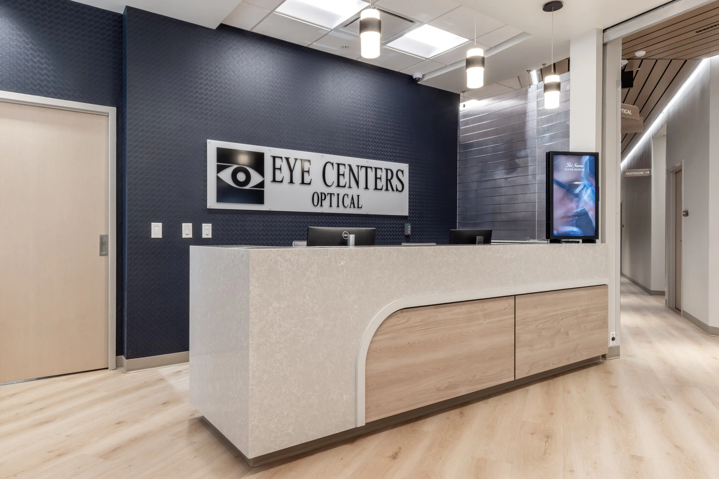 Reception desk at an eye care center with sign reading "Eye Centers Optical" on a dark textured wall, digital screen displaying a promotional image, and modern lighting fixtures in a clean, professional environment.