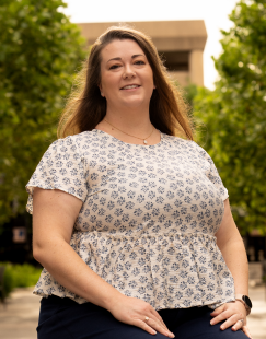 A woman smiling outdoors on a sunny day with trees and a building in the background.