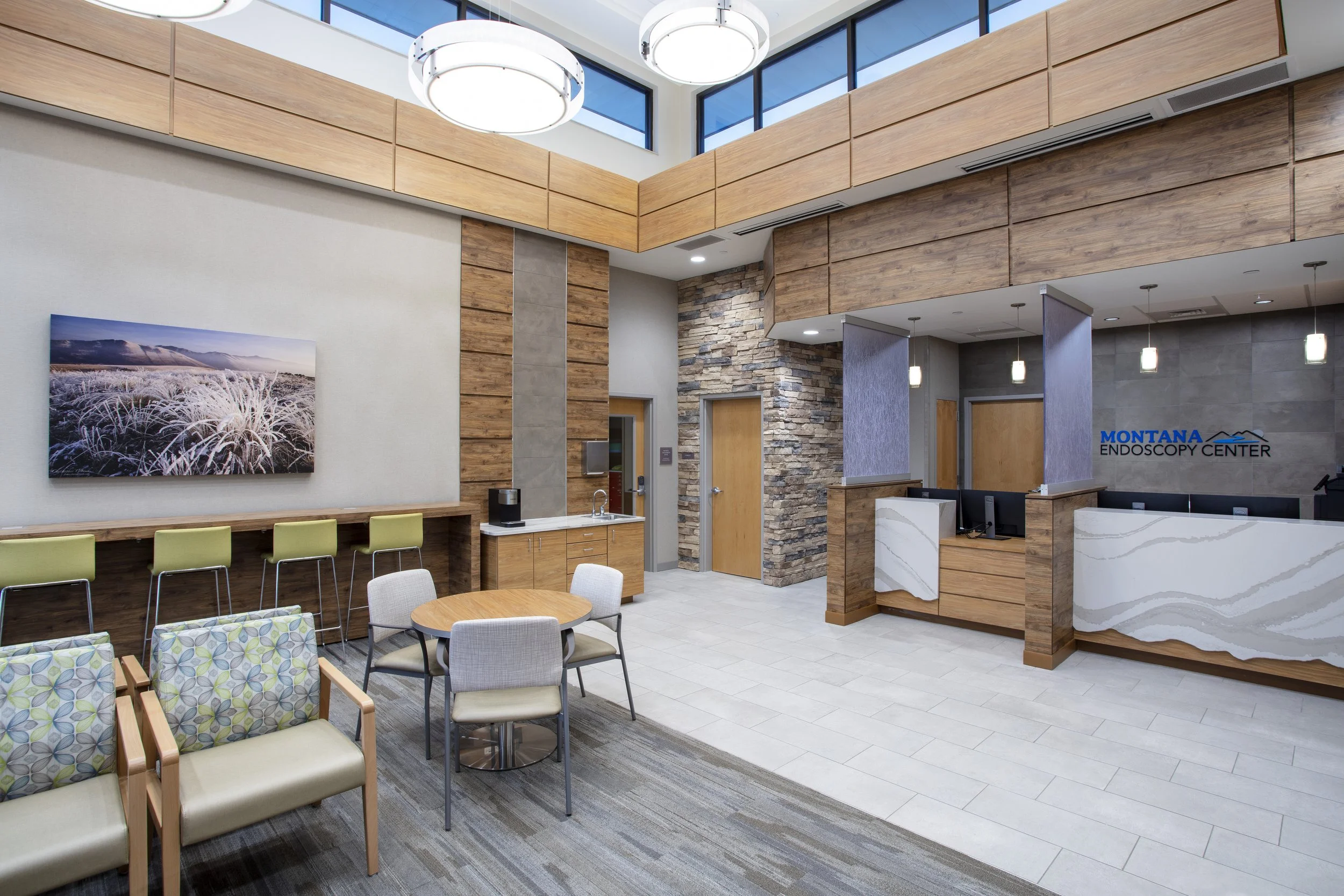 Interior of a modern medical office reception area with wooden accents, a stone wall, a front desk with a marble design, seating area with patterned chairs, and a large photograph of a frosty landscape on the wall.