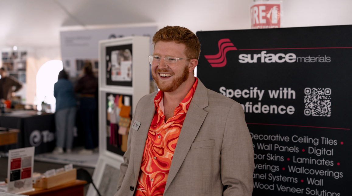 A smiling man with red hair, glasses, and a beard in a suit jacket and a vibrant orange patterned shirt standing in front of a display booth at a trade show or exhibition.