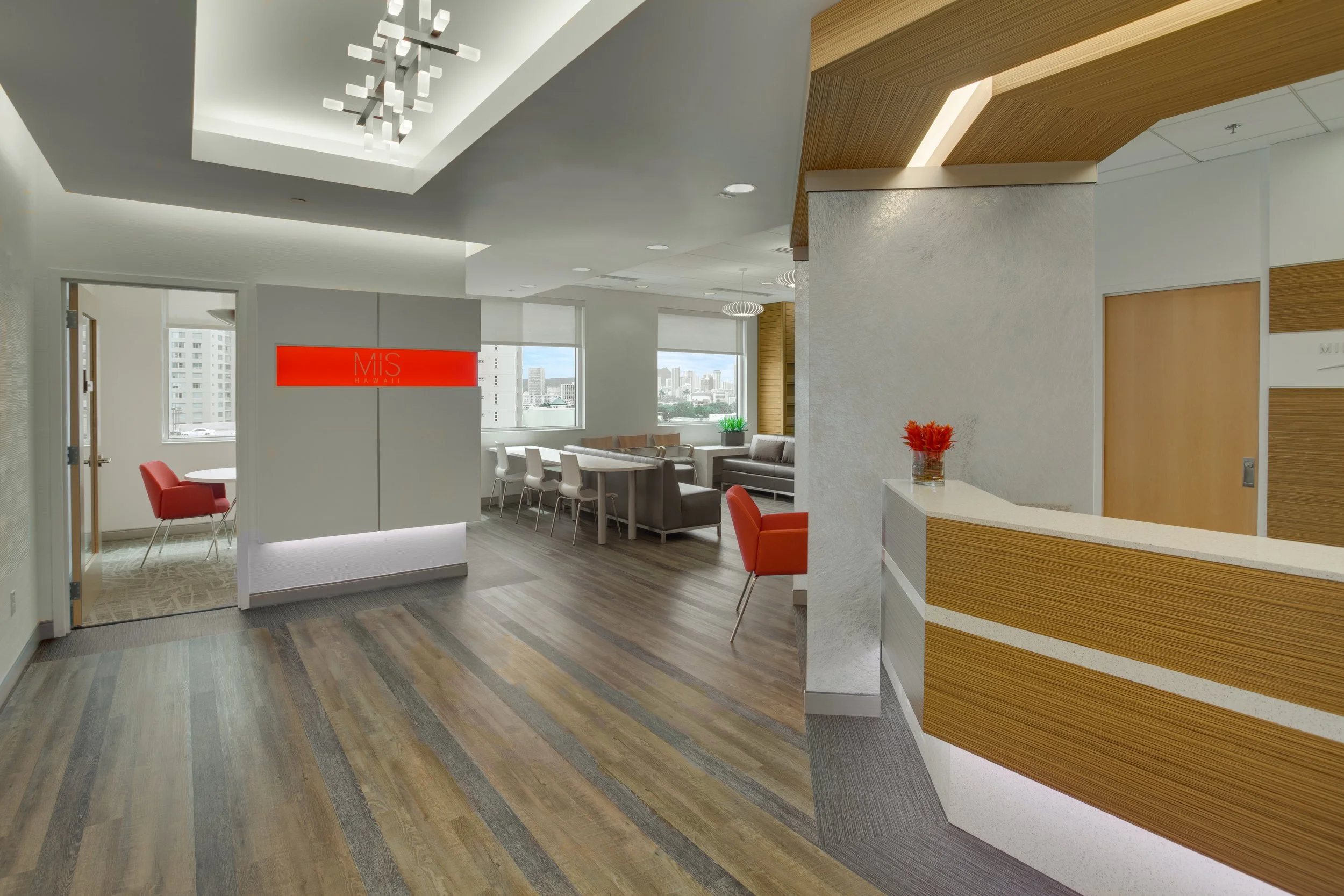 Modern office waiting area with white walls, natural wood accents, a staircase with wood paneling, multiple chairs, and large windows showing city buildings outside.