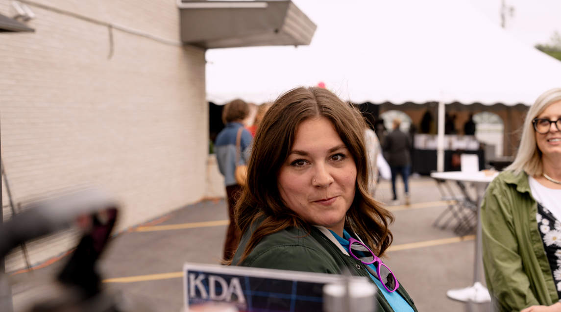 Young woman with brown hair and purple glasses hanging from her shirt collar, facing camera, with other people and a white tent in the background at an outdoor event.