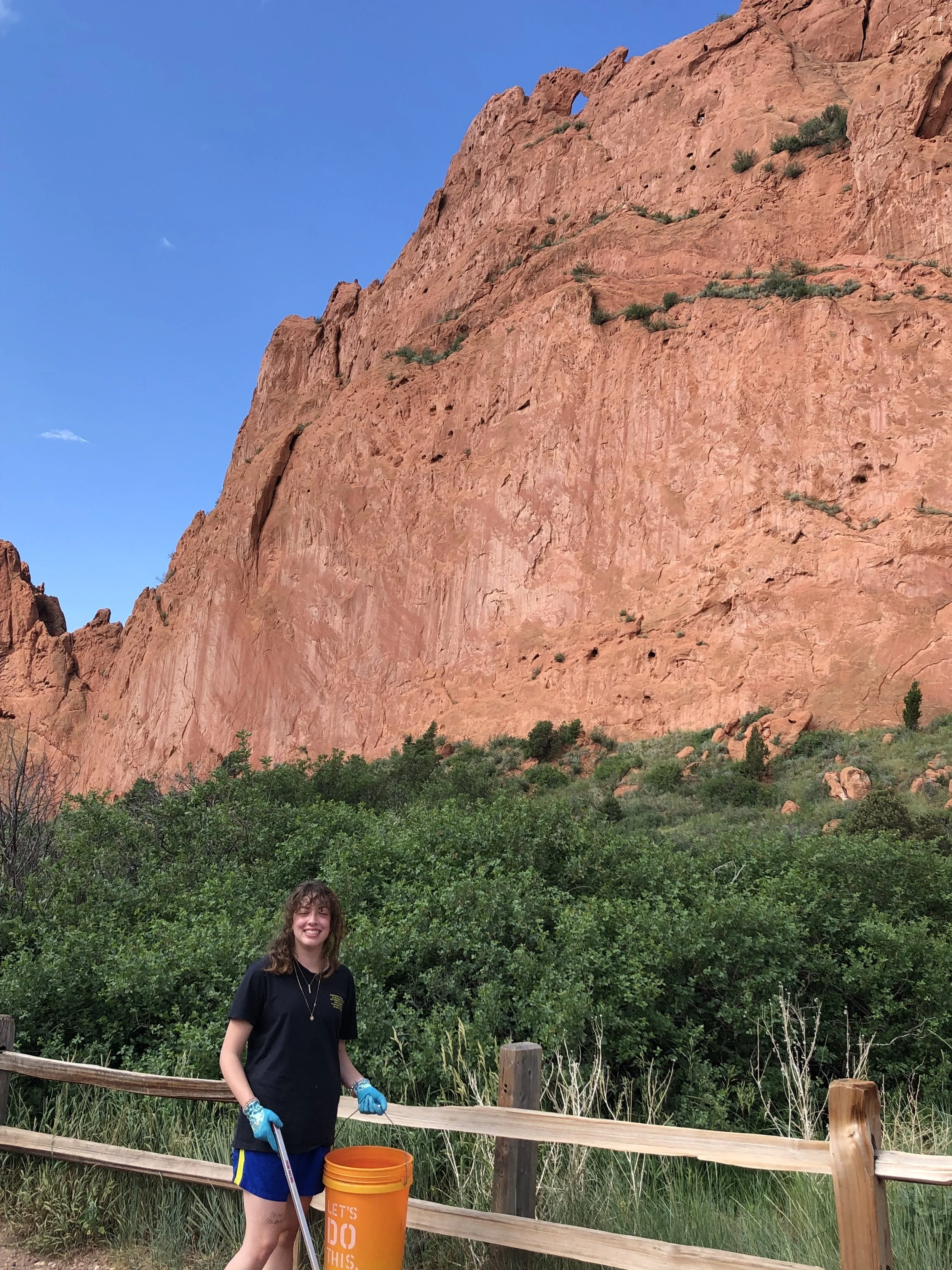 A young woman in black t-shirt, blue shorts, and blue gloves standing outdoors near a wooden fence, holding an orange trash bucket and trash picker, with a large red rock formation and green foliage in the background.