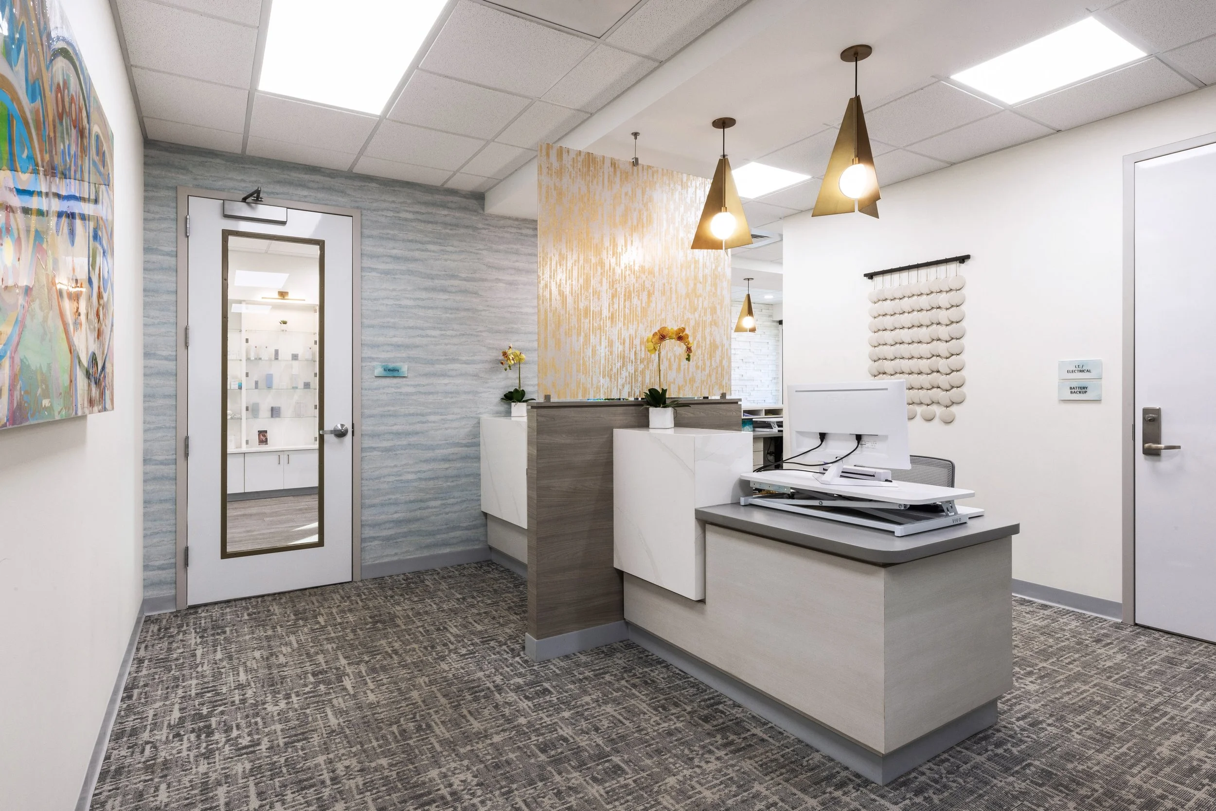 Modern office reception area with a curved white desk, decorative wall panels, hanging pendant lights, and artwork on the wall.