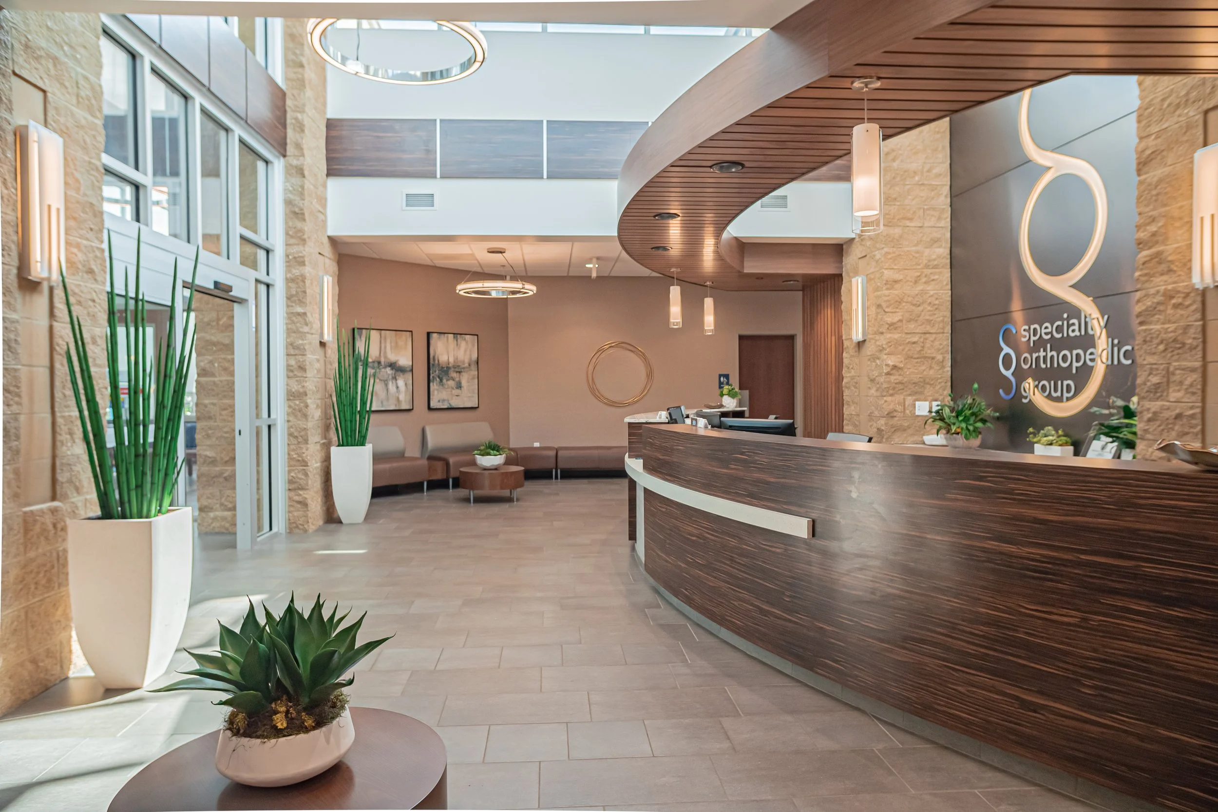 Interior of a medical office lobby with a curved wooden reception desk, beige stone walls, potted plants, seating area, and modern lighting fixtures.