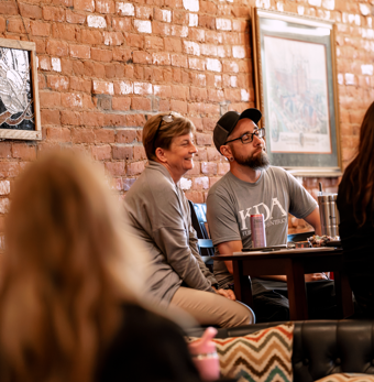 Two people sitting at a table in a coffee shop, smiling and talking, with a brick wall and framed pictures in the background.