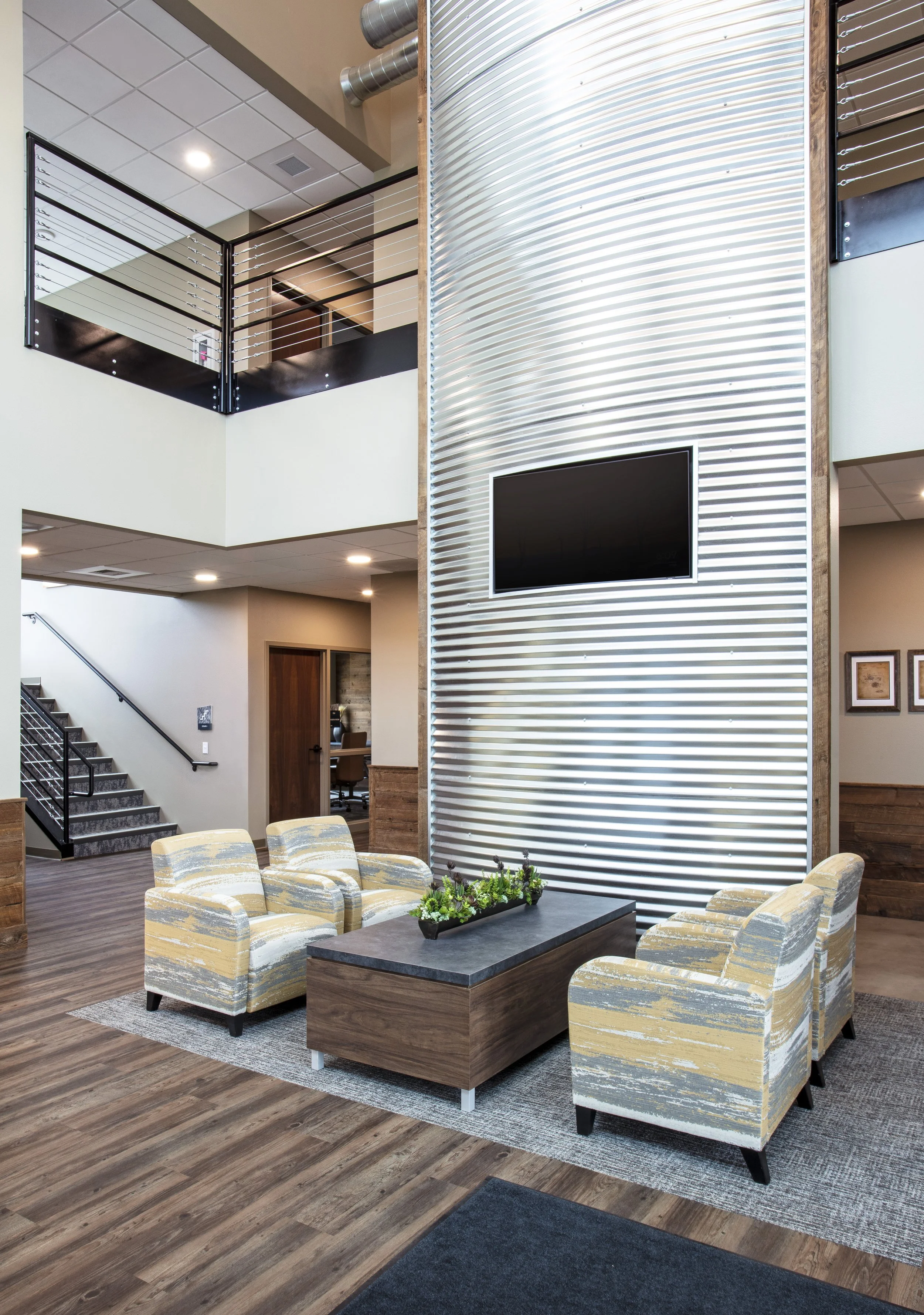 Lobby area with seating, a table with a plant, a large textured wall with a mounted TV, staircase, and second-floor balcony.