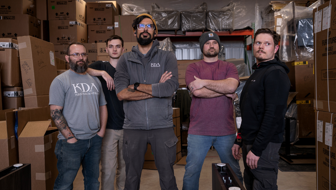 Five men standing in a warehouse surrounded by cardboard boxes and storage shelves, posing for a group photo.
