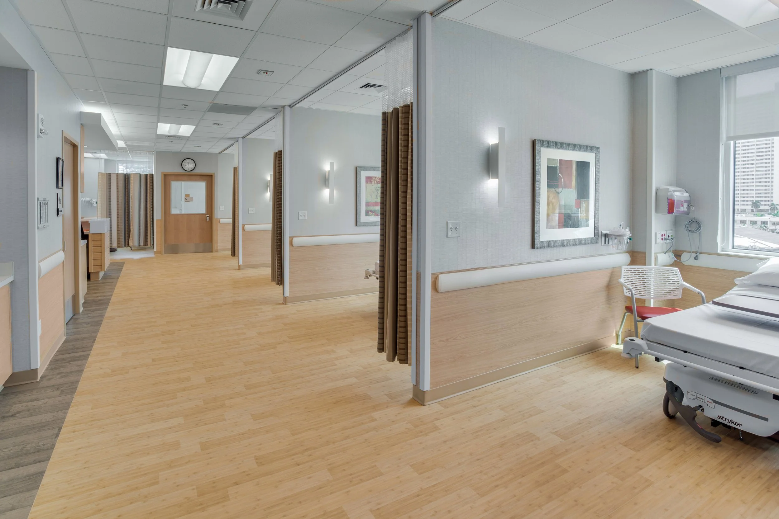 Hospital room with a hospital bed, medical equipment, and windows showing city buildings outside.