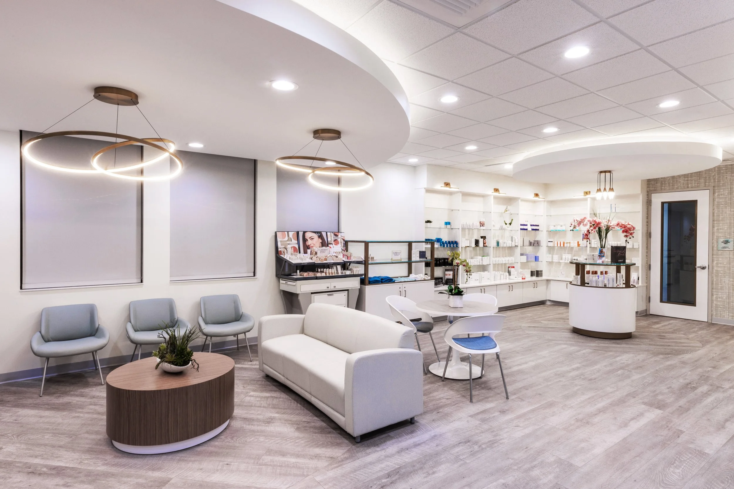 A modern waiting area with light gray chairs, a white sofa, a round wooden coffee table with a potted plant, and multiple decorative lighting fixtures on the ceiling. There are white shelves with various products on the right side, a round ceiling feature with spotlights, and windows with roller blinds.