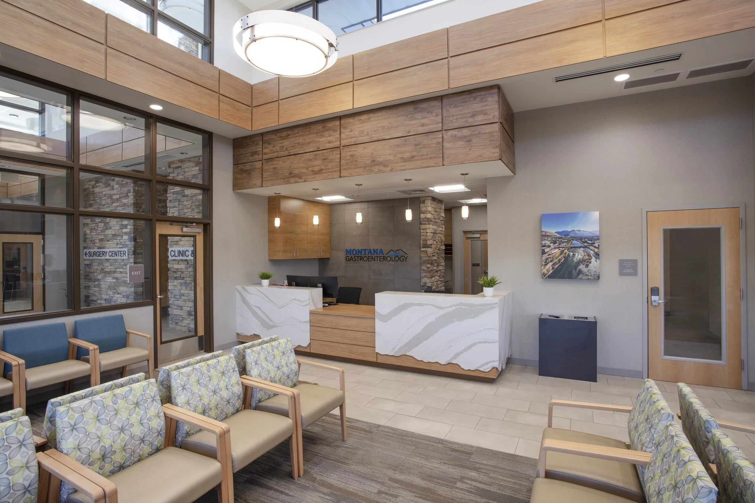 Interior of a medical clinic reception area with chairs, a reception desk, and signs indicating a surgery center and clinic. Modern decor with wood paneling, stone accents, and ceiling lights.