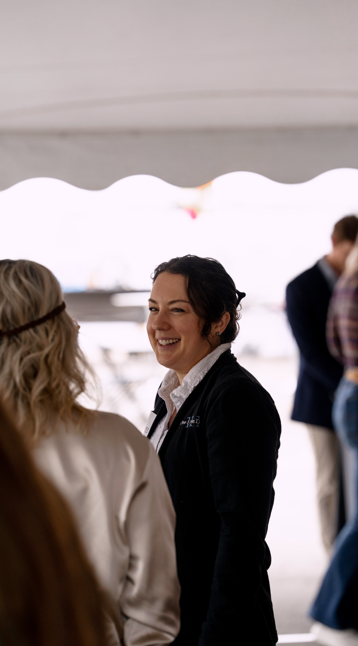 A woman smiling and talking to another person at an outdoor event, with a canopy overhead and boats in the background.