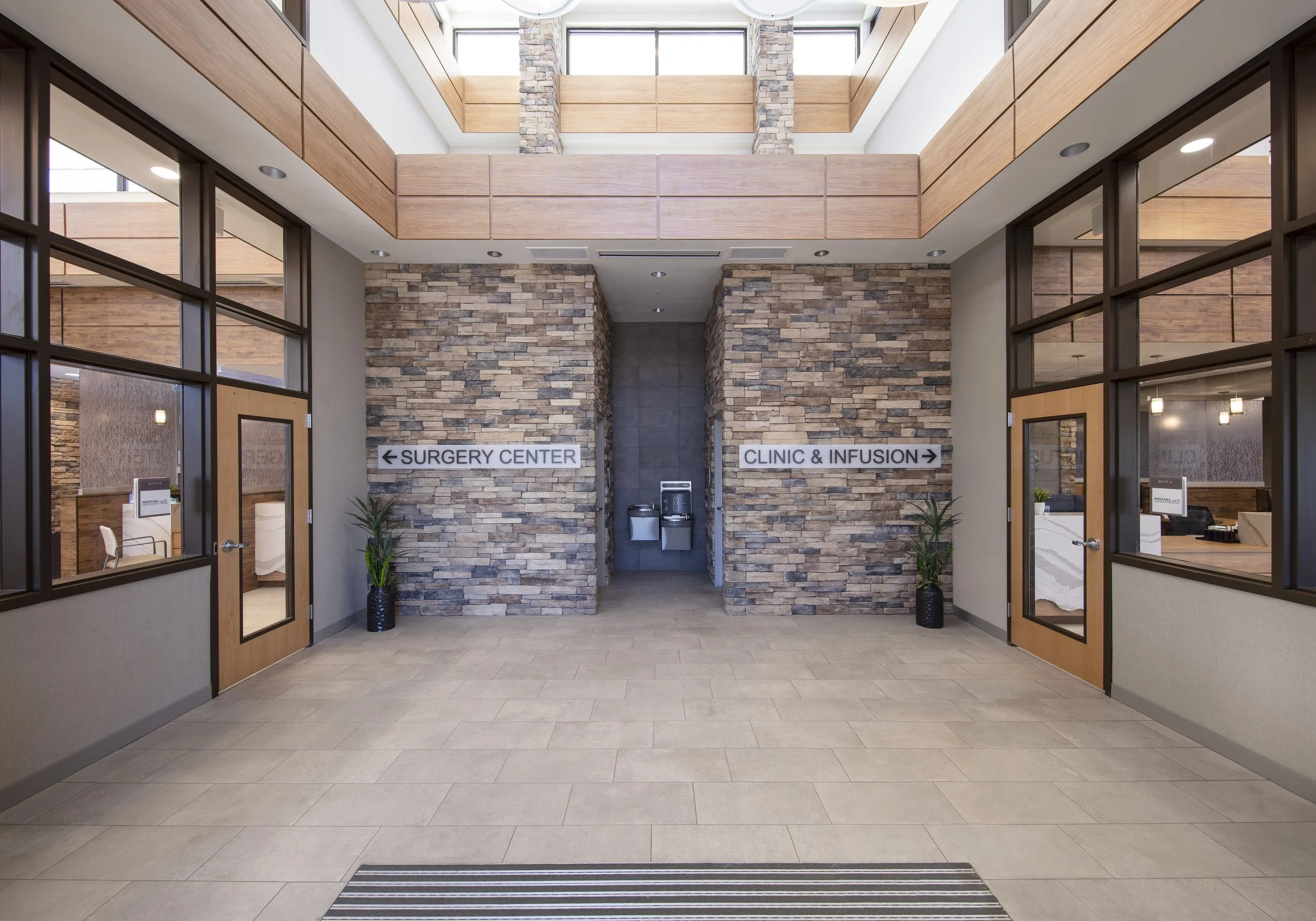 Interior view of a medical facility with brick walls, glass doors, and signs pointing to the surgery center on the left and clinic and infusion on the right.
