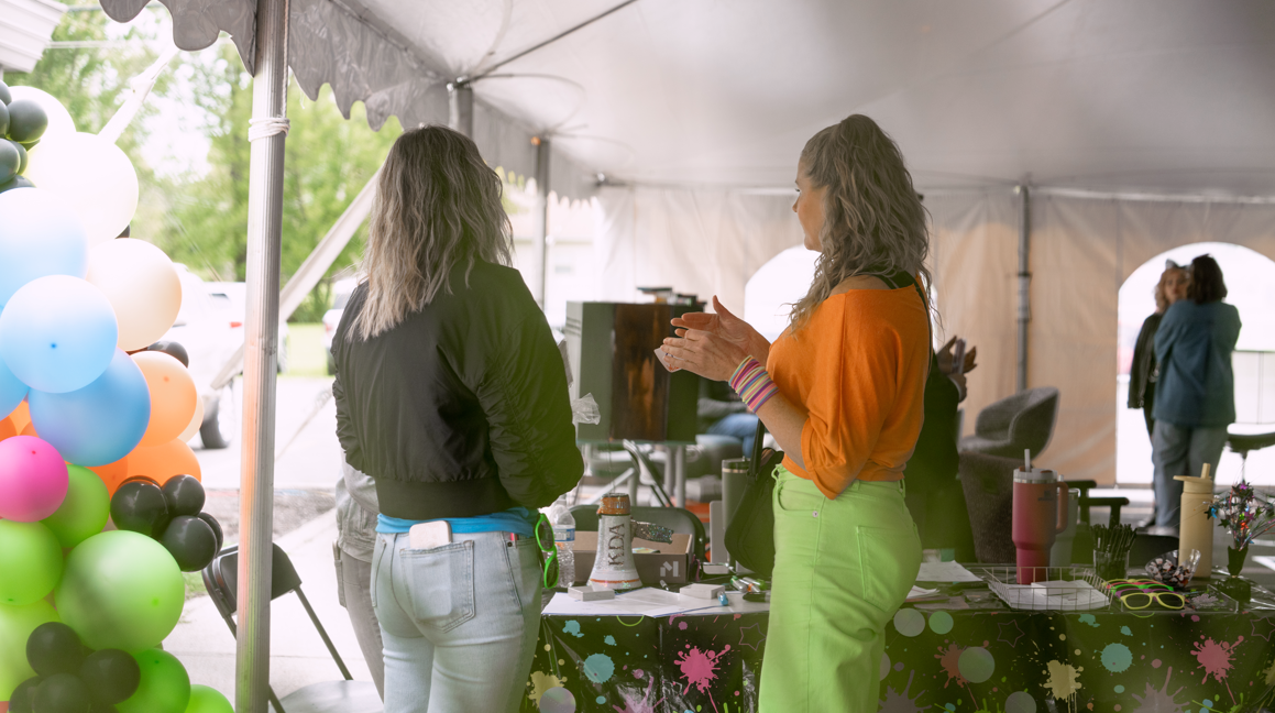 Two women talking inside a tent at an outdoor event, with colorful balloons on the left and a table with various items in front of them.