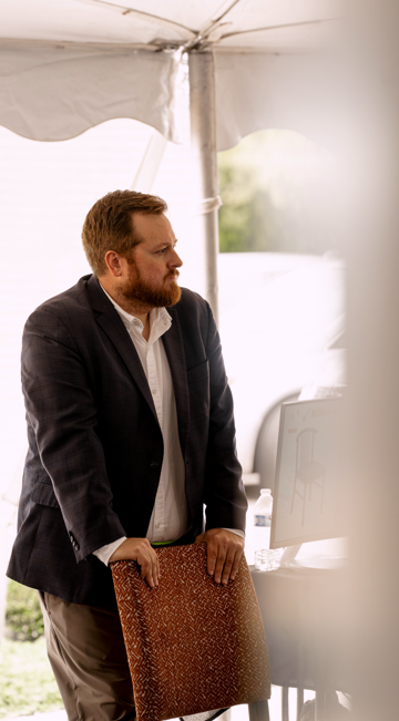 A man with a beard and brown hair, dressed in a dark blazer, standing behind a chair in front of a computer inside a tent.