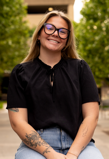 A smiling woman with long blonde hair wearing glasses, a black top, and jeans, sitting outdoors with green trees in the background.