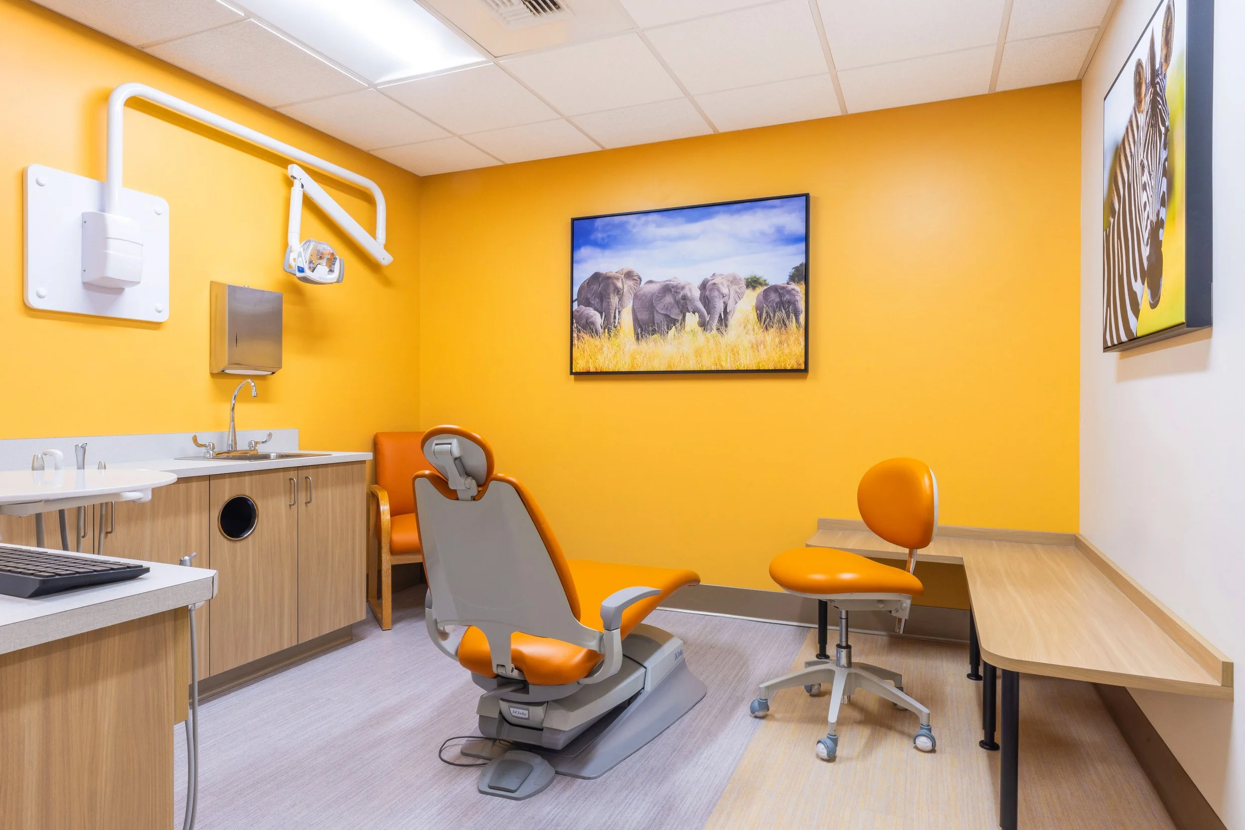 A medical examination room with yellow walls, featuring an orange examination chair, a framed picture of elephants, a sink, and various medical equipment.