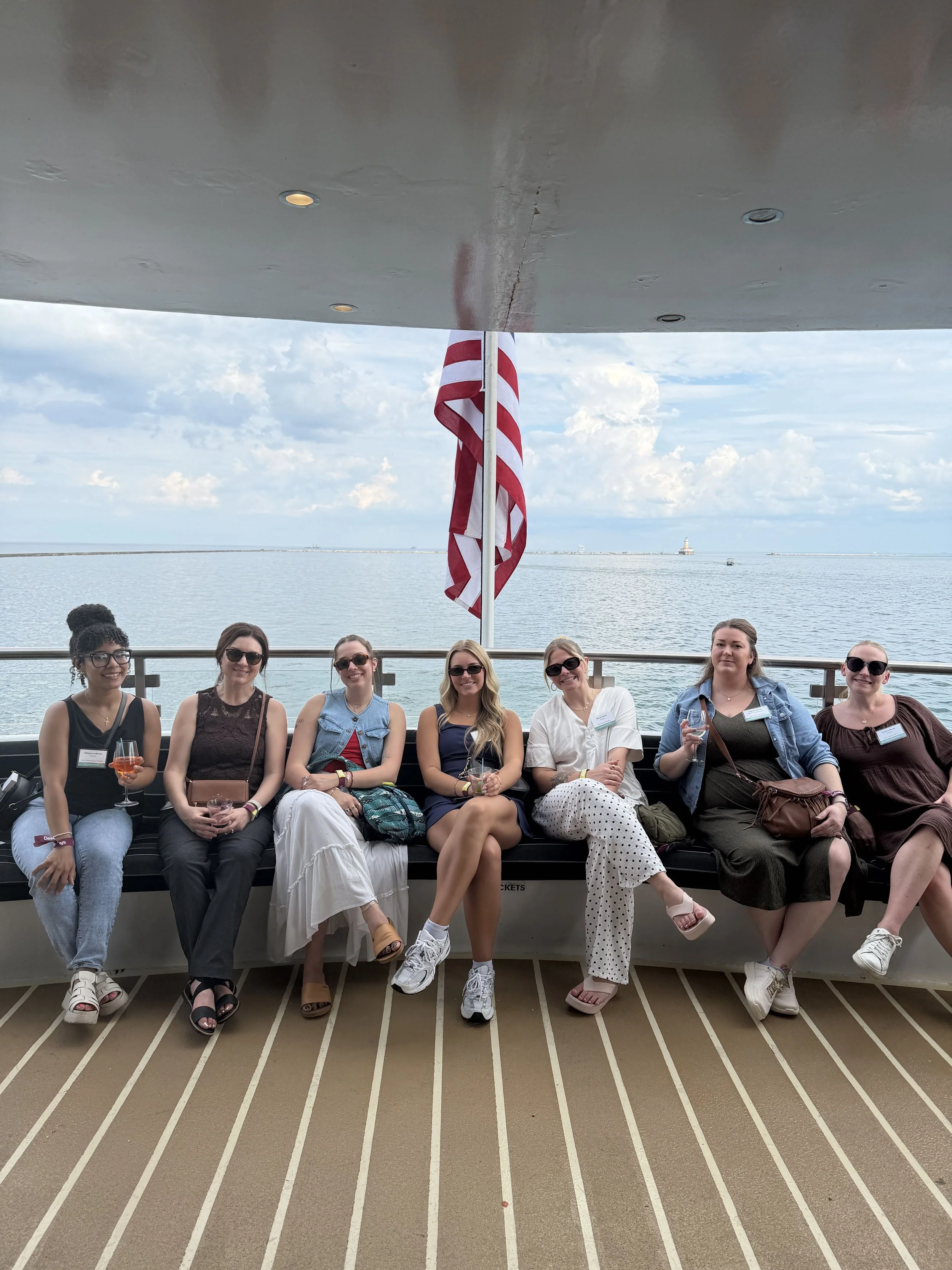A group of seven women sitting on a boat with the ocean and lighthouse in the background. They are smiling, holding drinks, and wearing sunglasses.