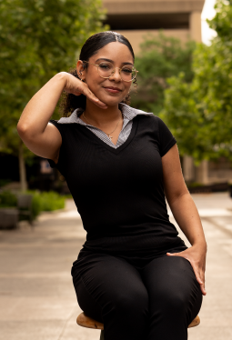 Young woman with glasses and dark hair smiling, sitting outdoors in a black shirt, with trees and a building in the background.