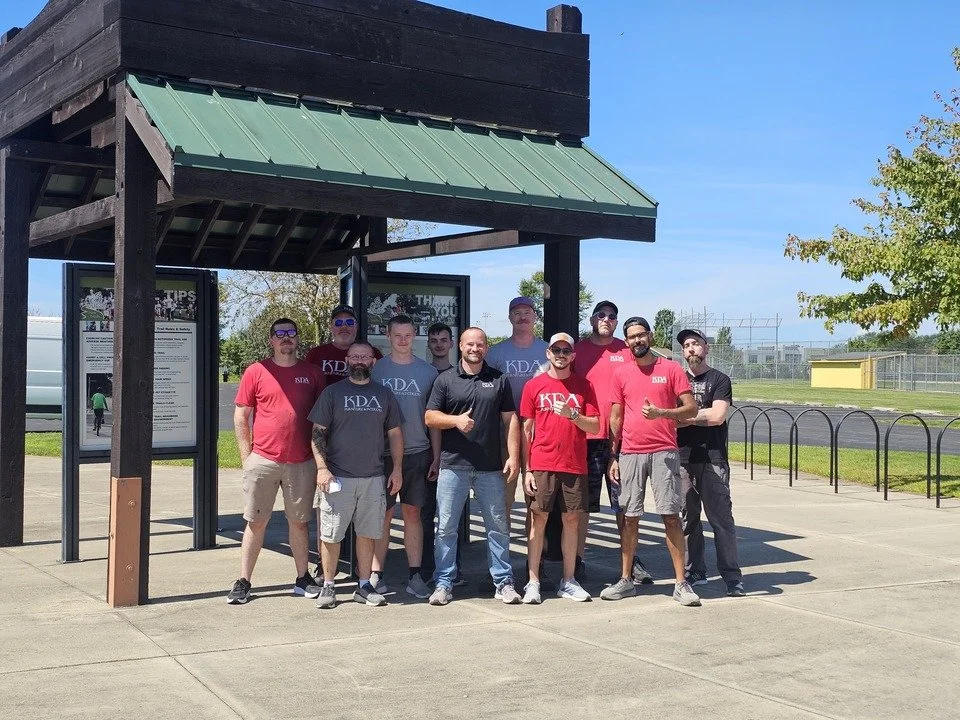 Group of men standing outdoors under a bus stop shelter, some wearing KDA shirts, posing for a photo on a sunny day.