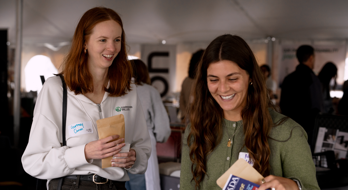 Two women smiling and looking at a pamphlet or flyer at a professional event or conference.
