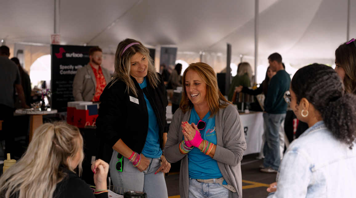 Two women engaging in conversation at an event, smiling, surrounded by other attendees, with booths and tables in the background.