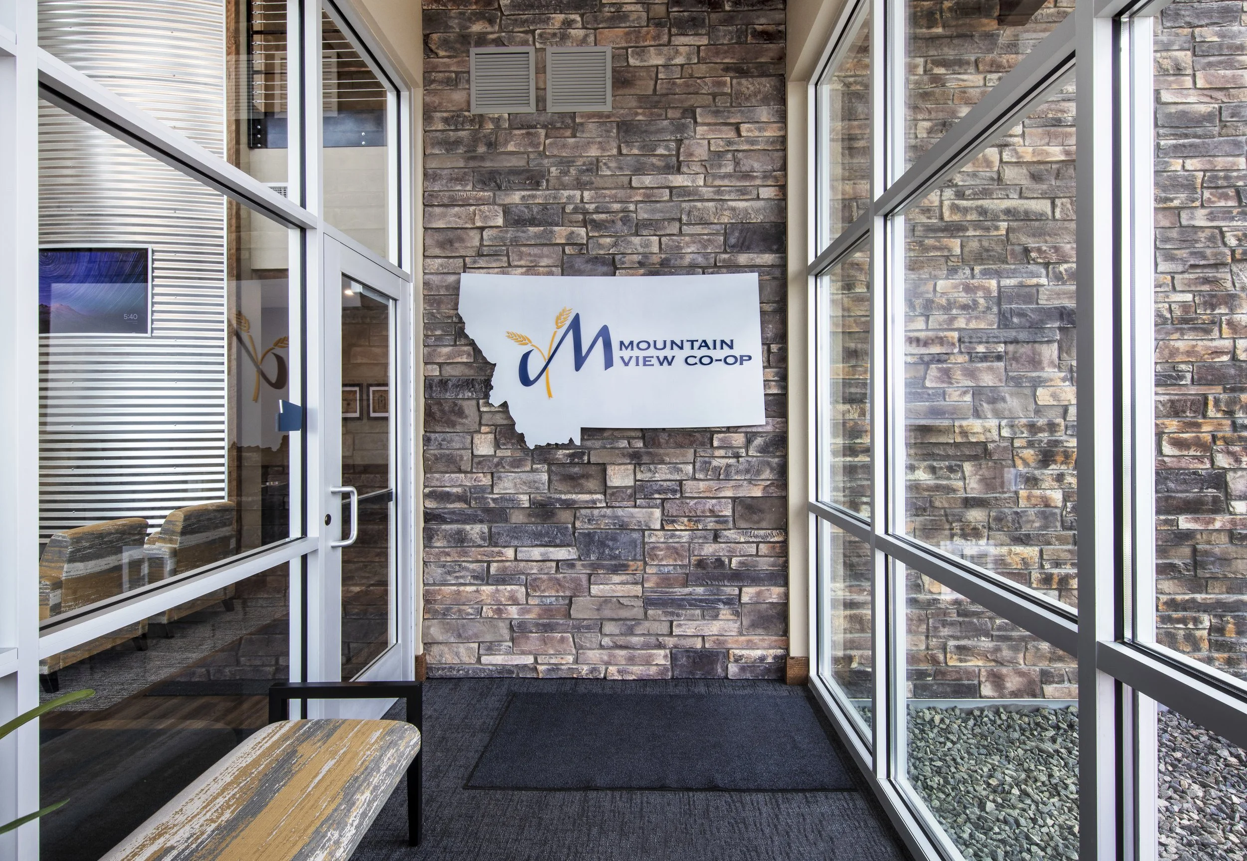Entrance of the Mountain View Co-op with glass windows and a brick wall, a sign with the organization's name and logo, and indoor seating with wooden benches.